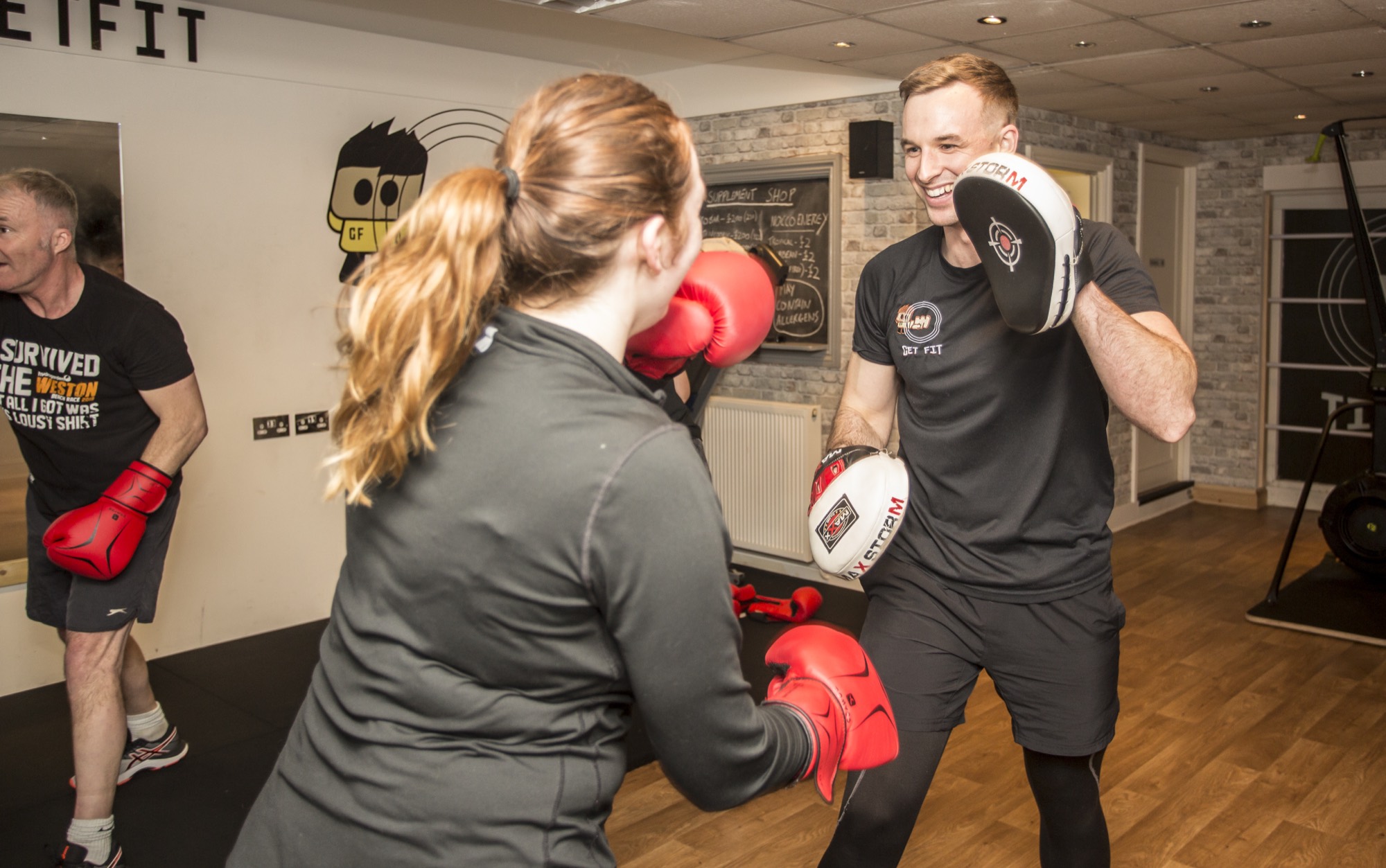 Inside Get Fit Studio: Matt in black holding focus pads while a client in red boxing gloves throws a punch; the Get Fit cartoon mascot painted on the wall behind