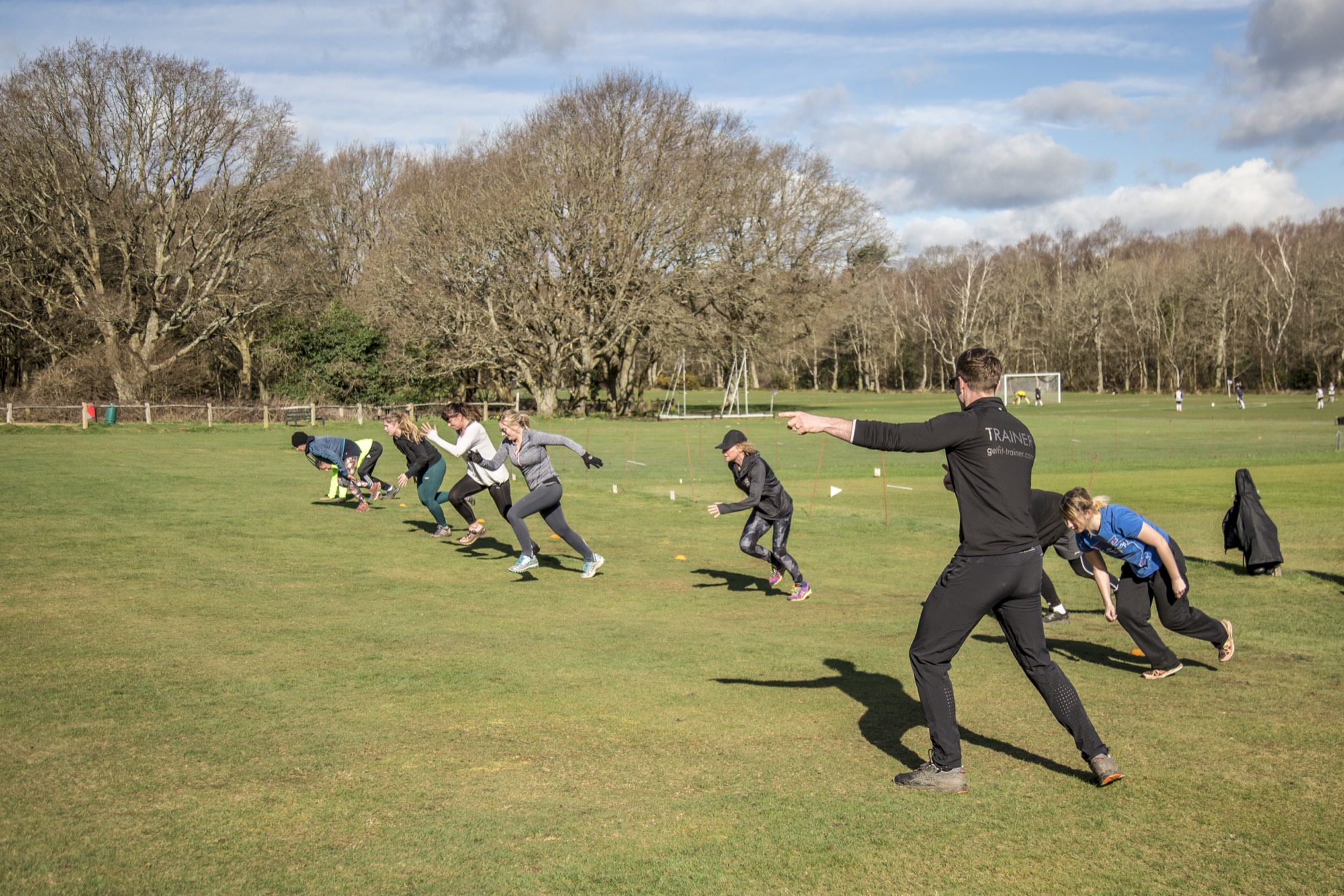 Sunday outdoor bootcamp on a Henfield playing field: a line of about ten people mid-sprint with Matt out front, pointing the way, bare winter trees behind