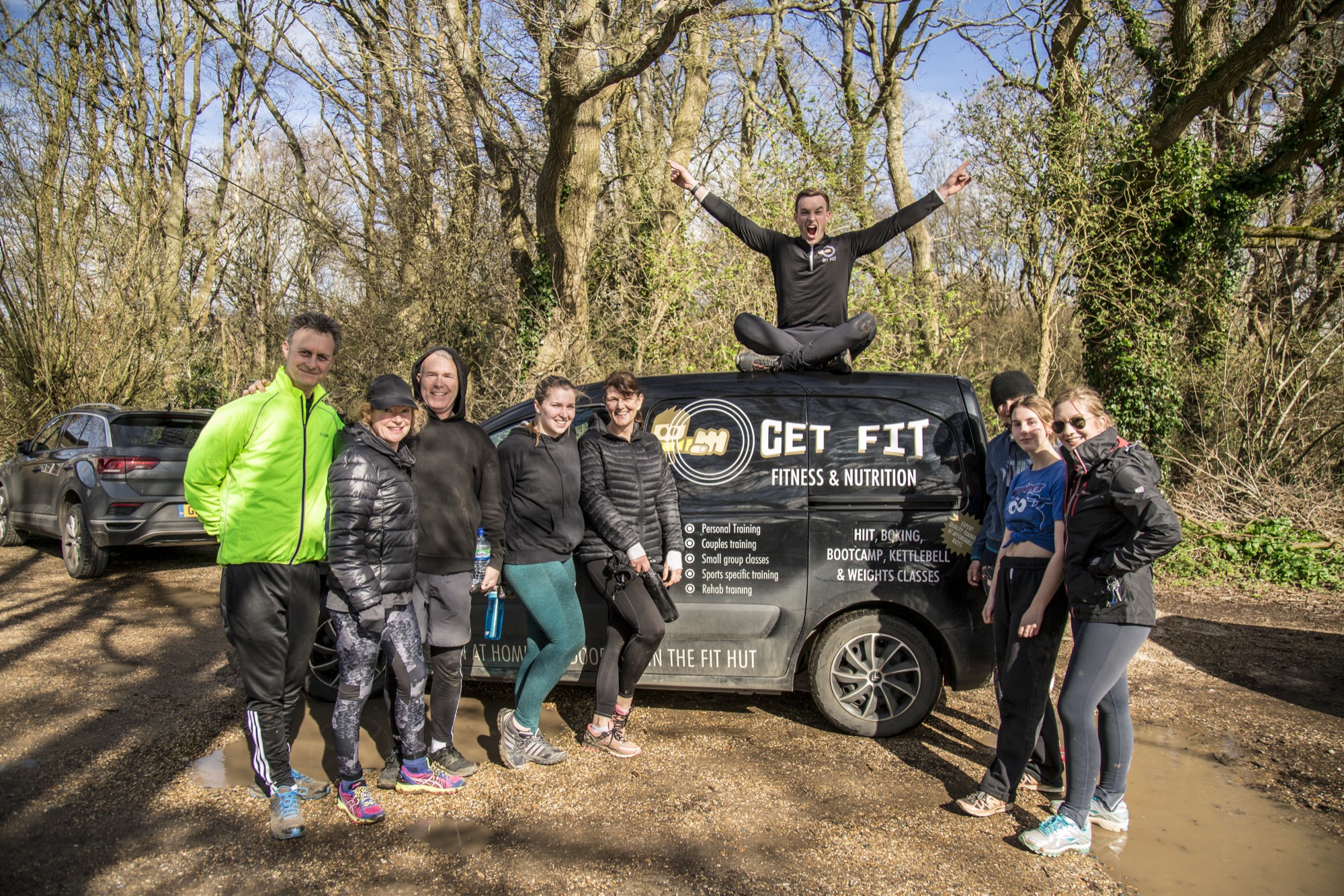 Ten clients lined up beside the black Get Fit van, one man leaping in the air on its roof, arms up, on a woodland track